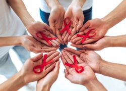 geöffnete Hände, im Kreis angeordnet, halten eine rote AIDS-Schleife