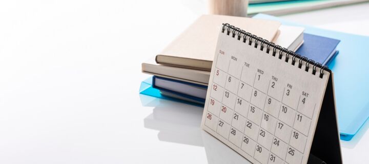 stehender Kalender mit Monatsansicht auf Schreibtsich mit Büchern und Stiftehalter im Hintergrund