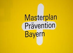 Schriftzug Masterplan Prävention Bayern auf gelbem Hintergrund mit weißem Plus hinterlegt