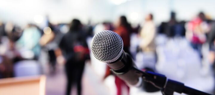 Ansprechpartner Nahaufnahme eines Mikrofons, stehende Personen und Plenum im Hintergrund