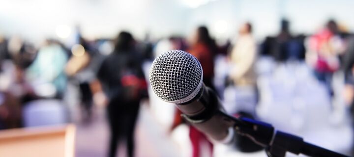 Nahaufnahme eines Mikrofons, stehende Personen und Plenum im Hintergrund