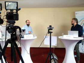 Aufbau der Online-Übertragung mit Stehtischen, Kameras, Roll-up, Referent, Moderator und Kameramann